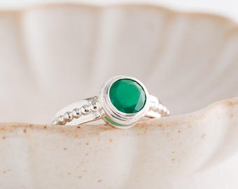 May include: A silver ring featuring a large, round, emerald-green gemstone. The ring has a bezel setting and a beaded band design. It rests in a light-coloured, scalloped-edge ceramic dish. The ring is a piece of jewellery.