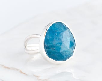 May include: A silver ring featuring a faceted, blue gemstone. The gemstone is teardrop-shaped and set in a silver band. The ring sits on a white, textured surface. The blue stone is the main feature.