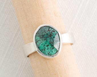 May include: A silver ring with a large, oval turquoise stone. The stone has a mottled green and black pattern. The ring band is a simple, flat design. The ring is displayed on a wooden dowel.