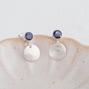 Peut inclure: Une paire de boucles d'oreilles pendantes en argent avec une pierre précieuse ronde et facettée bleu foncé sertie dans une monture en argent, reliée à un petit disque en argent texturé et circulaire suspendu sous la pierre.