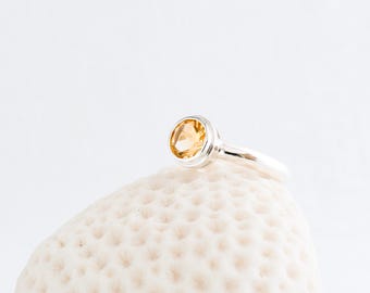 May include: A silver ring featuring a round, light orange gemstone. The ring is displayed on a white, textured surface. The gemstone is set in a silver bezel. The band is smooth and polished, reflecting light.