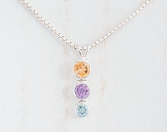 May include: A silver pendant necklace with three gemstones: orange, purple, and light blue. The gemstones are set in silver bezels and hang from a fine silver chain. The necklace is a delicate piece of jewellery.