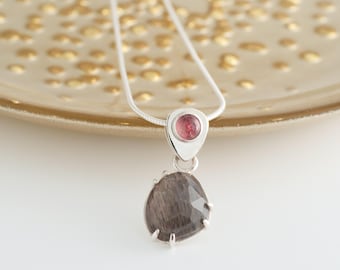May include: A silver pendant necklace with a teardrop-shaped, faceted, dark grey gemstone and a small, round, pink gemstone. The pendant hangs from a delicate silver chain. The necklace is displayed on a gold-coloured surface, showcasing a minimalist design.