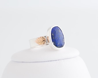 May include: A silver ring with a large, oval, deep blue lapis lazuli stone. The ring has a gold-coloured leaf detail on the band. The gemstone is set in a silver bezel, creating a striking contrast.