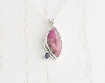 May include: A silver pendant necklace with a large, oval pink gemstone and a small blue gemstone. The pendant is suspended from a fine silver chain, offering a simple yet elegant design. The pink gemstone is approximately 3 cm in length.