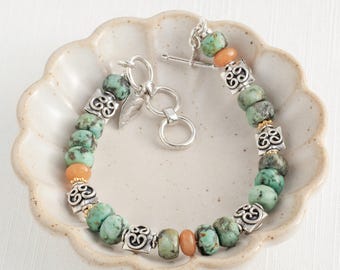 May include: A bracelet with turquoise and peach-coloured beads, accented by silver-toned metal spacers. The bracelet, secured with a toggle clasp, is presented in a decorative, off-white ceramic dish. The beads are approximately 0.8 cm in diameter.