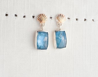 May include: Silver earrings featuring rectangular, faceted, light blue gemstones. Each earring has a gold seashell design at the top. The earrings are presented on a white, textured background.
