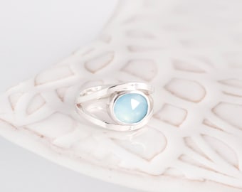 May include: A silver ring featuring a light blue, oval gemstone. The ring has a split band design, with the gemstone set in the centre. The ring is displayed on a white, textured surface.