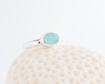 May include: A silver ring featuring a light blue oval gemstone. The ring is displayed on a textured, off-white surface. The band is simple, drawing attention to the polished gemstone. The ring's design is elegant, emphasizing the stone's colour and shape.