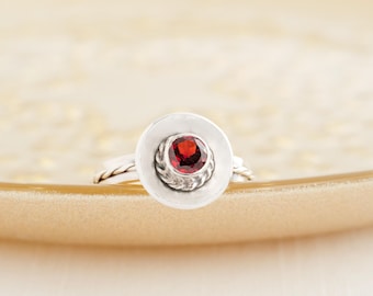 May include: A silver ring with a central, deep red gemstone set within a circular frame. The gemstone is encircled by a braided metal design, and the ring has a plain band. The ring is presented on a light-toned surface.