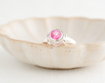 May include: A silver ring featuring a large, round, bright pink gemstone. The ring is set in a simple, elegant design and is placed in a cream-coloured, scalloped-edge ceramic dish. The ring is the focal point, showcasing the vibrant pink stone.