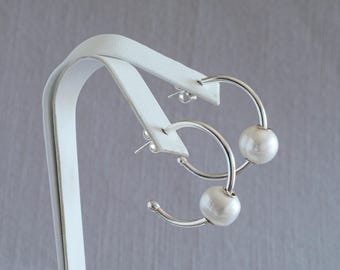 May include: Silver hoop earrings featuring large, round, white pearl details. The earrings are presented on a white stand against a grey backdrop. The design of the earrings is simple and elegant.