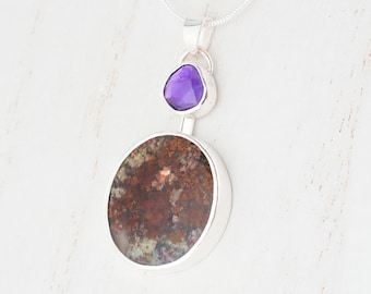 May include: A silver pendant necklace. It features a large, round, speckled brown and green stone, and a smaller, purple, teardrop-shaped stone. The stones are set in silver and suspended from a delicate silver chain.