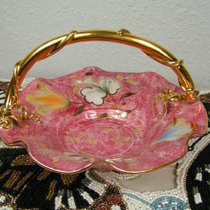 May include: Pink ceramic basket with a gold handle and rim. The basket features floral designs in white, orange, and gold. The handle is a twisted gold design. The basket is likely for decorative purposes.