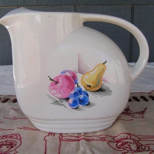 May include: White ceramic pitcher with a handle, featuring a painted design of a pear, apple, and grapes. The pitcher has a rounded body and a spout.