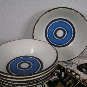 May include: A set of ceramic bowls and a plate. The bowls and plate are white with a blue and white circular design in the center. The rims are dark brown. The bowls are stacked, and the plate is beside them.