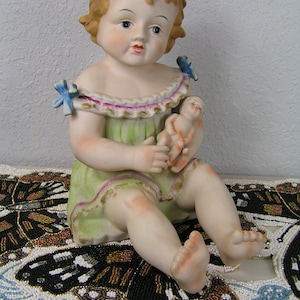 May include: A porcelain figurine of a seated child doll, wearing a green dress with pink trim and blue accents. The doll holds a smaller doll. The figurine is on a beaded surface with a butterfly design.