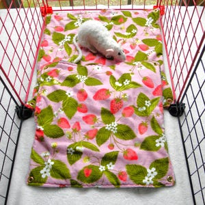 May include: A pink and green strawberry-patterned pet bed inside a red wire cage. The bed has a matching pillow and a white stuffed rat toy. The bed is designed for small pets.