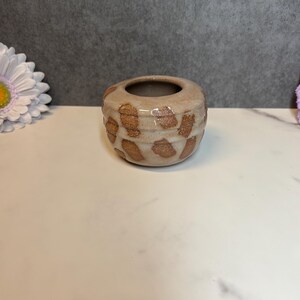 May include: A small, round ceramic vase with a speckled pattern in shades of tan and brown. The vase has a dark brown interior and a textured exterior. The design is reminiscent of a giraffe's spots. The vase is suitable for small flowers or decorative purposes.
