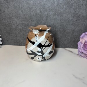 May include: A small, hand-painted ceramic vase with a unique design. The vase has a brown base with white and black paint splatters. The rim has a wavy, textured edge. This decorative vase is suitable for small floral arrangements.