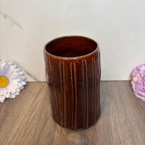 May include: A handcrafted, cylindrical ceramic vase with a rich, dark brown glaze. The vase features vertical, textured lines, giving it a rustic appearance. The open top reveals the interior. This decorative piece is suitable for home decor.