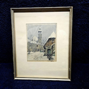 May include: Framed print of a winter scene, showing a snow-covered town with a church and buildings. The artwork is in a silver frame. The print depicts a snowy street with bare trees and a church steeple.