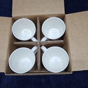 May include: Four white ceramic coffee cups with handles, neatly arranged inside a cardboard box with dividers. The cups are empty and have a glossy finish, suggesting they are new. The box is open, revealing the contents.