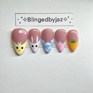 May include: Set of five almond-shaped press-on nails with spring-themed designs. Each nail features a different design, including a chick, bunny, clouds, flowers, and a carrot. The nails have a pink base with white, yellow, blue, and orange accents. The brand name "Blingedbyjaz" is in a black rectangle.