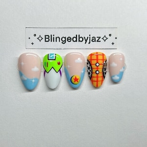 May include: A set of five press-on nails featuring custom designs including a space ranger suit, a yellow plaid pattern, a colorful ball, and blue sky with white clouds. The text Blingedbyjaz is printed on a white rectangular label above the nails.