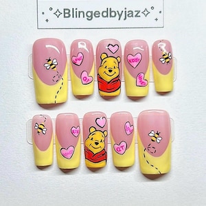 May include: Set of ten artificial nails with a yellow and pink design. Each nail features a cartoon image of Winnie the Pooh, hearts, and bees. The text "Blingedbyjaz" is at the top.