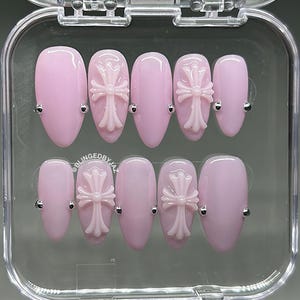 May include: A set of ten almond-shaped press-on nails in a soft pastel pink finish. Four nails feature a raised, light pink cross emblem, and all nails are accented with small silver-toned metal studs. The text BLINGEDBYJAZ is printed on one nail.