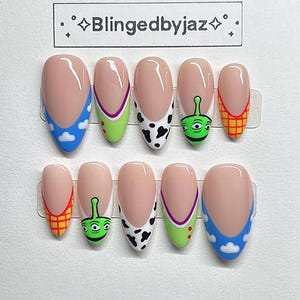 Puede incluir: Un conjunto de diez uñas postizas con diseños inspirados en la película Toy Story. Los diseños incluyen cielos azules con nubes, Buzz Lightyear, un estampado de vaca, un alien verde y el chaleco de Woody. El texto "Blingedbyjaz" está en la parte superior.