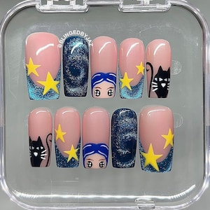 Puede incluir: Un juego de diez uñas postizas con base nude, puntas de purpurina azul, estrellas amarillas, ilustraciones de gatos negros y rostros con cabello azul. El texto BLINGEDBYJAZ está impreso en la uña superior dentro de un estuche transparente.