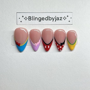 May include: Set of five almond-shaped press-on nails with a nude base and colorful designs. The designs include a blue nail with a red bow, a lavender nail with a pink accent, a red nail with black accents, a red polka dot nail, and a yellow nail with green accents. The text "Blingedbyjaz" is above the nails.