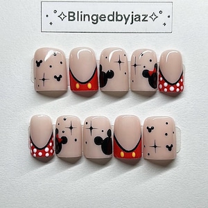 May include: A set of ten press-on nails with a nude base, decorated with Mickey Mouse designs. Some nails feature black Mickey Mouse silhouettes, starbursts, and red and white polka dot accents. The brand name "Blingedbyjaz" is printed on the top.