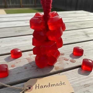 Może przedstawiać: Grupa czerwonych, sześciennych przedmiotów zawieszonych na czerwonym sznurku, z kilkoma luźnymi kostkami rozrzuconymi wokół. Dołączona jest brązowa etykieta z napisem "Handmade". Przedmioty znajdują się na drewnianej powierzchni.