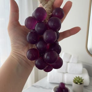 以下が含まれることがあります： 手で持たれた、自然な麻ひものループが付いた紫色のブドウの房。ブドウは深く、半透明な色をしています。背景には、白いタオルと小さな多肉植物のあるバスルームの洗面化粧台が含まれています。