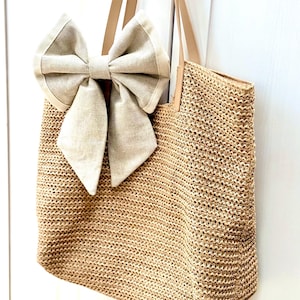 May include: A woven straw tote bag with a large beige bow attached. The bag has light brown leather handles and a rectangular shape. The bow is made of a textured fabric and adds a decorative element to the bag.