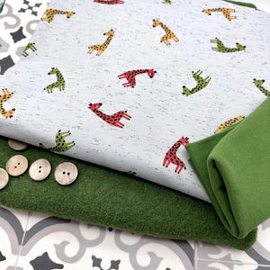 Könnte beinhalten: Stapel von Stoffen mit einem Giraffenmuster in Rot, Gelb und Grün auf einem gesprenkelten weißen Hintergrund. Ein grüner Stoff und Holzknöpfe sind ebenfalls zu sehen. Geeignet für Näh- oder Bastelprojekte.