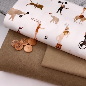 Könnte beinhalten: Stoffstapel mit Zirkusmotiven mit braunen und beigen Artisten und Tieren. Holzknöpfe mit Blattmustern liegen neben dem Stoff. Der Stoff ist auf einer braunen Oberfläche geschichtet.