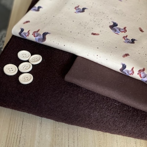 Peut inclure: Gros plan sur deux échantillons de tissu. L'échantillon du haut est un tissu crème avec un motif répétitif d'écureuils et de feuilles brunes. L'échantillon du bas est un tissu polaire brun foncé. Il y a cinq boutons blancs sur le tissu.