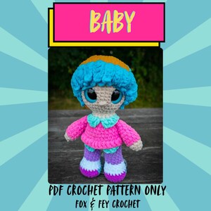 Op de afbeelding: Een handgemaakte gehaakte pop met blauw en geel haar, grote blauwe ogen en een roze outfit. De pop draagt een roze trui met een lichtblauwe kraag, een paarse broek en gestreepte schoenen. Het woord "BABY" staat in een roze banner.