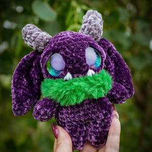 May include: A handmade purple crochet monster plushie with grey horns and large, iridescent eyes. It has a green furry collar and a soft, textured appearance. The plushie is designed with large ears and a friendly expression.