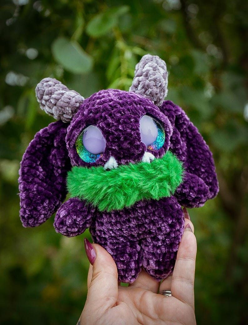 May include: A handmade purple crochet monster plushie with grey horns and large, iridescent eyes. It has a green furry collar and a soft, textured appearance. The plushie is designed with large ears and a friendly expression.