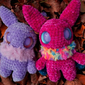 May include: Two crocheted plush toys with large eyes. One is purple with a pink fur collar and the other is pink with a rainbow fur collar. The toys are sitting on a bed of brown leaves.