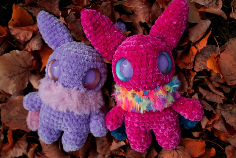 May include: Two crocheted plush toys with large eyes. One is purple with a pink fur collar and the other is pink with a rainbow fur collar. The toys are sitting on a bed of brown leaves.