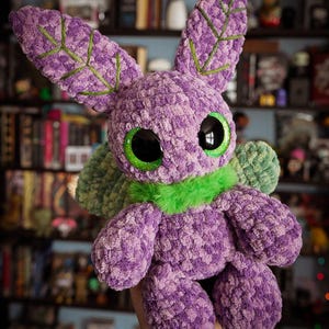 Puede incluir: Un juguete de peluche hecho a mano en tonos morado, verde y negro. El juguete tiene ojos verdes grandes y brillantes, orejas largas con detalles bordados en verde y un cuello de piel verde. El peluche se sostiene frente a un fondo borroso de libros y estanter&iacute;as.