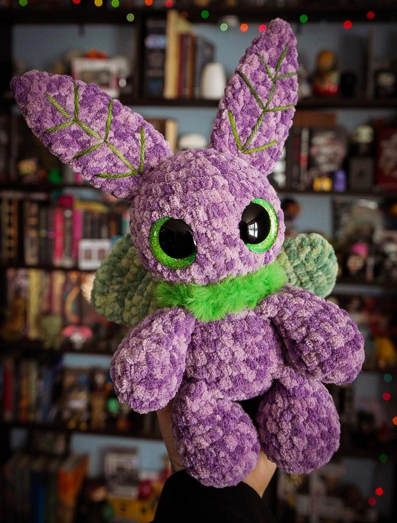 Puede incluir: Un juguete de peluche hecho a mano en tonos morado, verde y negro. El juguete tiene ojos verdes grandes y brillantes, orejas largas con detalles bordados en verde y un cuello de piel verde. El peluche se sostiene frente a un fondo borroso de libros y estanter&iacute;as.