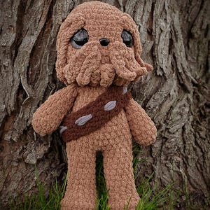 May include: A handmade brown crochet plush toy resembling a character with a furry appearance, large black eyes, and a bandolier. The toy is standing in front of a tree trunk and green grass.