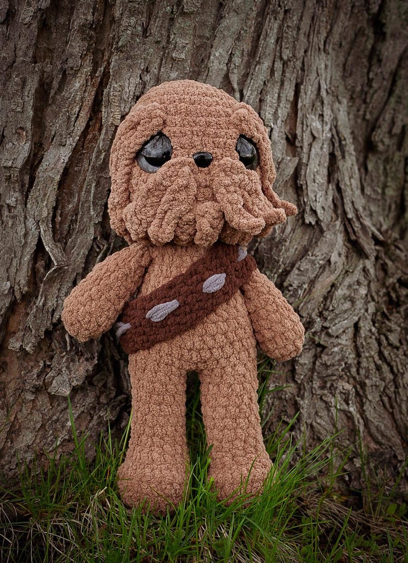 May include: A handmade brown crochet plush toy resembling a character with a furry appearance, large black eyes, and a bandolier. The toy is standing in front of a tree trunk and green grass.